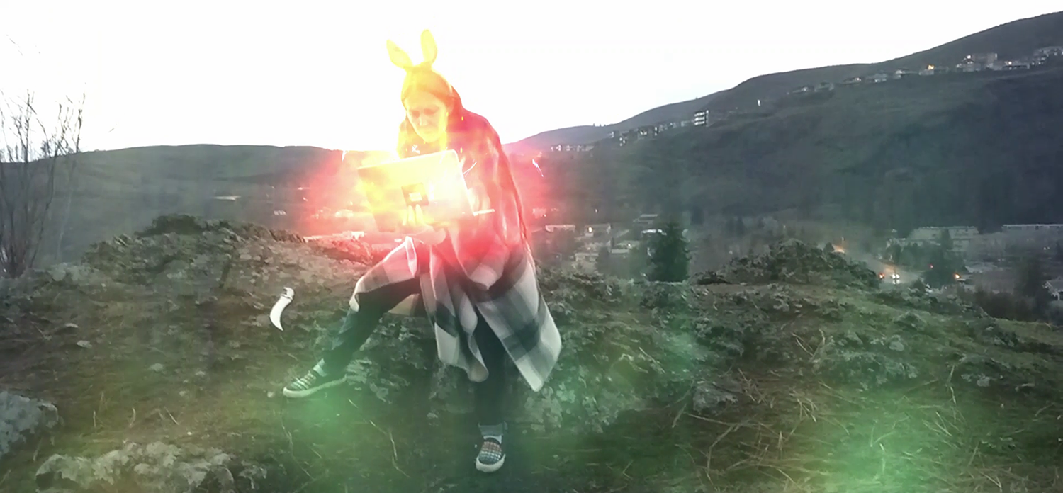 A woman in bunny ears types frantically on a glowing magical laptop on the top of a hillside