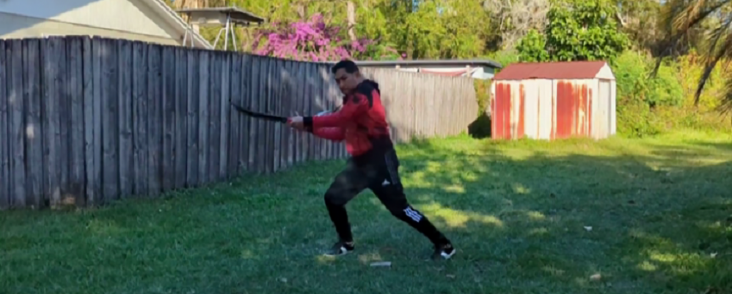 A man displaying his dual-weapon swordfighting in a warm green background that is obviously somebody's backyard