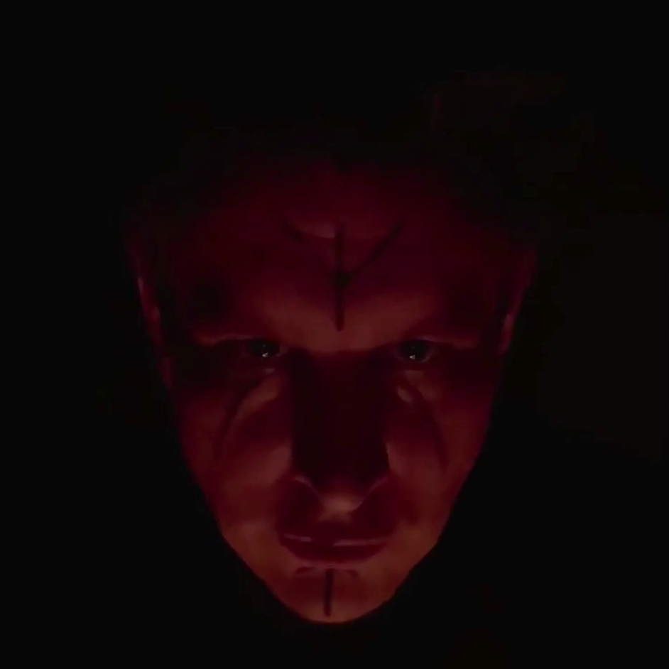 A man shrouded in shadow and firelight leaning close to the camera with runes painted on his face