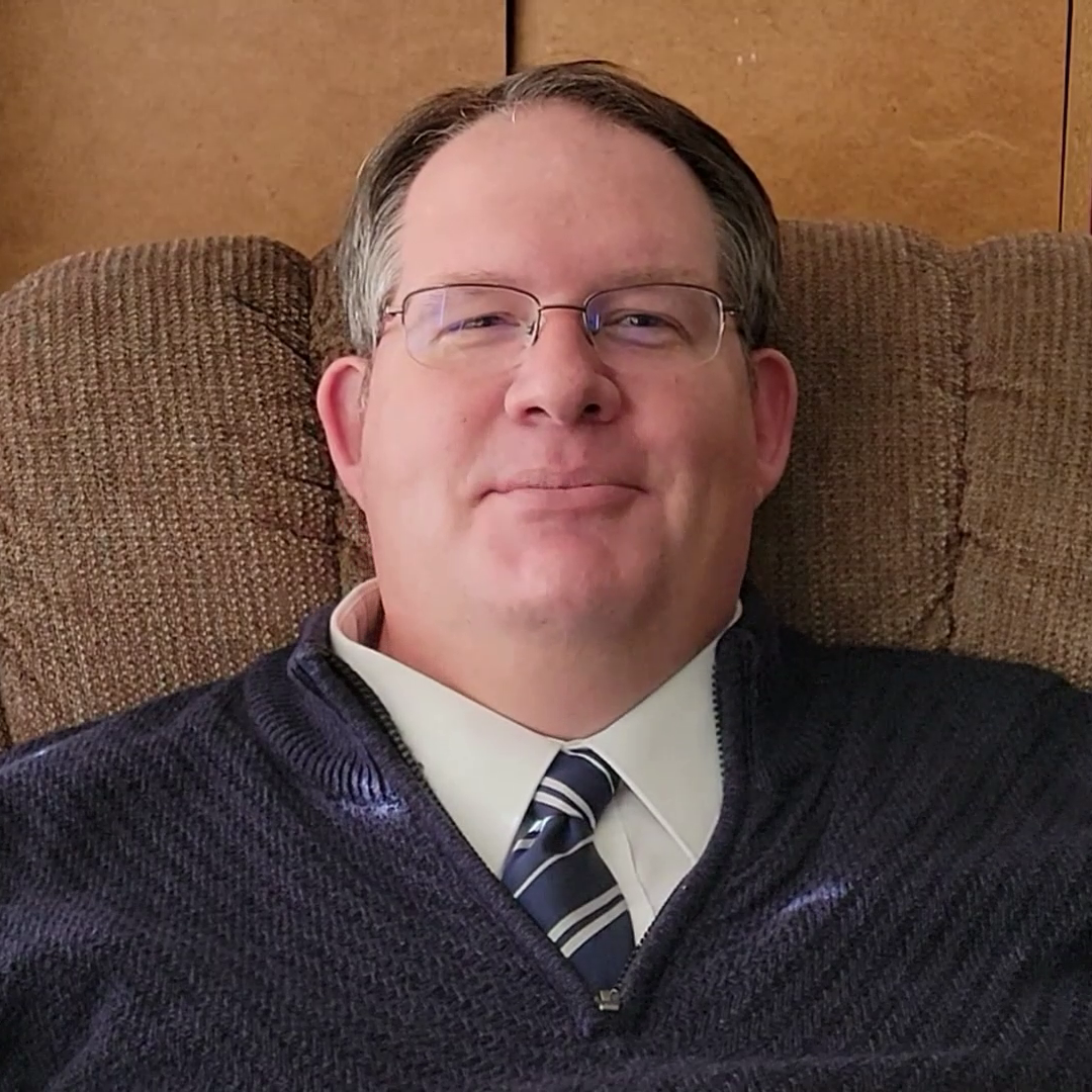 A smiling man in a sweater and tie sitting in an easy chair