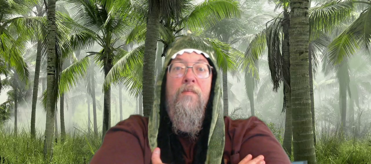A grey-bearded man in a dinosaur hat, with arms folded, in a jungle